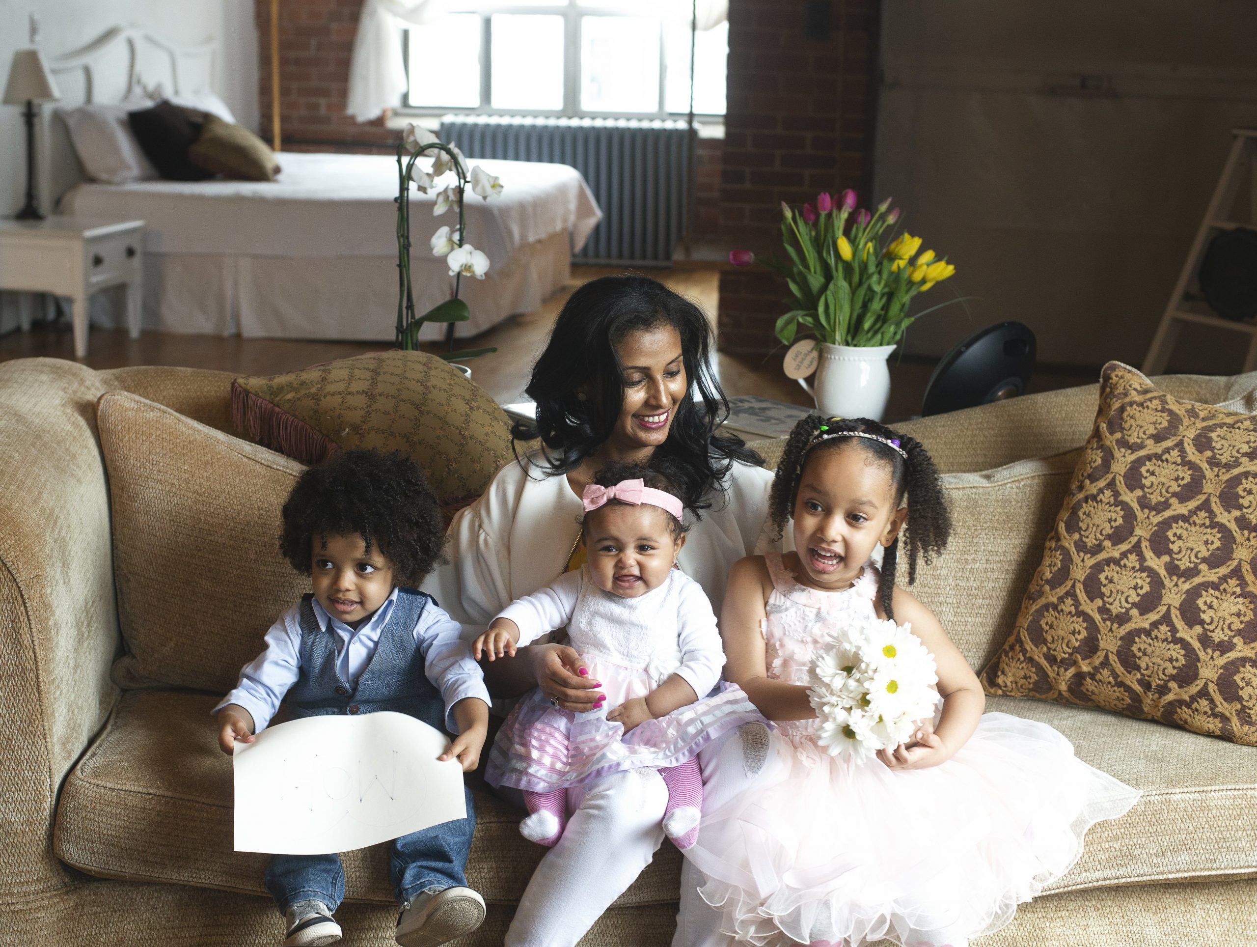 Mother with three young children, a young toddler in a suit on the left, a baby with a pink bow in the middle of the mother's lap, and a young girl in a pink dress holding flowers on the right.
